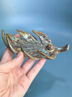 A hand holding a small ceramic fish figurine with a glossy, mottled glaze in shades of green, brown, and gold, set against a soft blue gradient background.