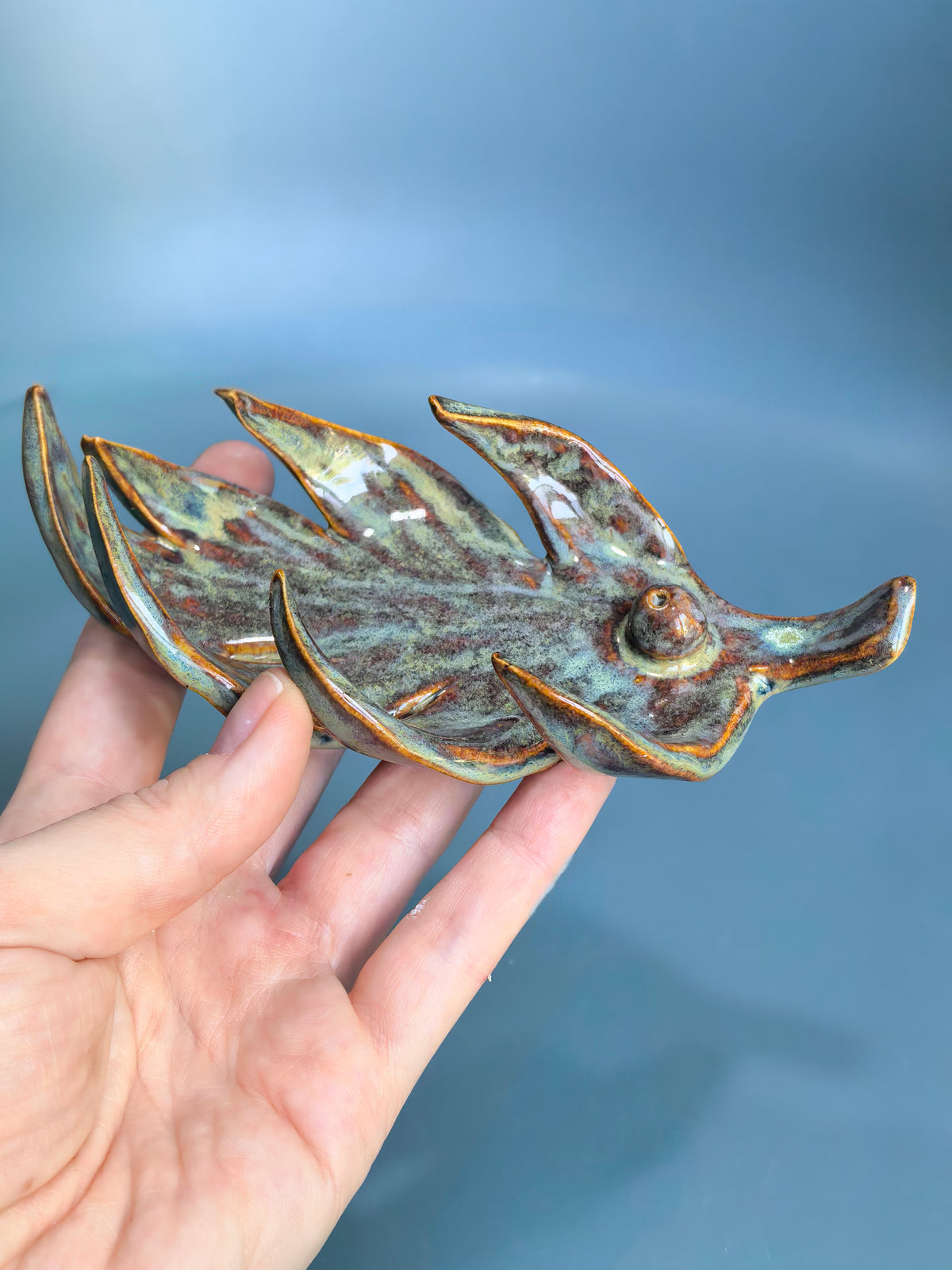 A hand holding a small ceramic fish figurine with a glossy, mottled glaze in shades of green, brown, and gold, set against a soft blue gradient background.