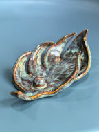 A ceramic leaf-shaped dish with a textured, organic shape and a glossy glaze featuring green, brown, and copper-orange tones, set against a smooth blue-gray background.