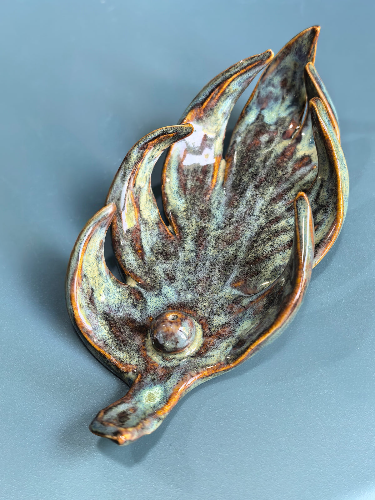 A sculptural ceramic dish shaped like a stylized leaf or flower bud, featuring a glossy glaze with mottled brown, olive green, and cream tones, set against a solid slate-blue background.