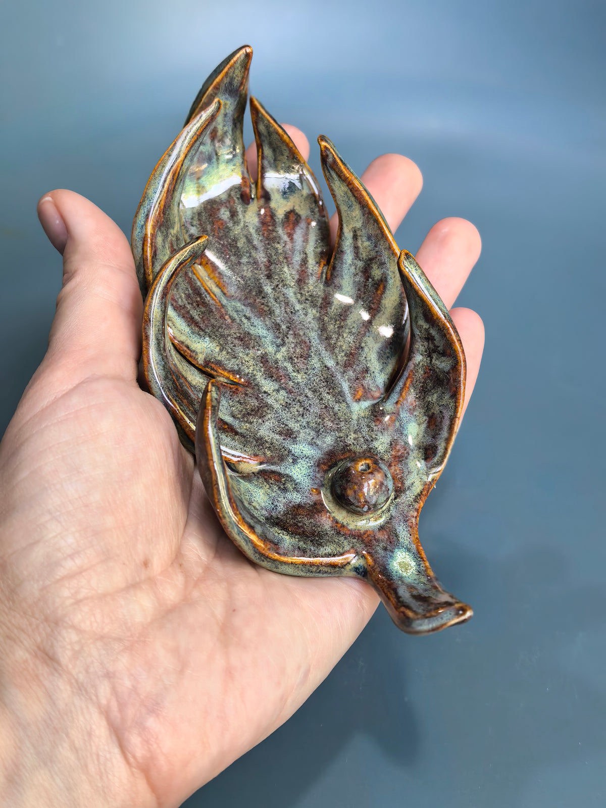 A hand holding a decorative ceramic dish shaped like a stylized leaf, featuring a glossy glaze with brown, green, and blue tones against a blue-gray background.
