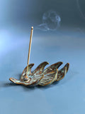 A ceramic incense holder shaped like a leaf with a single incense stick standing inside, photographed on a smooth blue studio background with soft smoke rising.