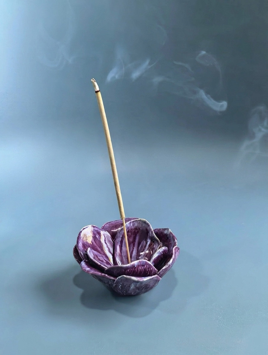 A purple ceramic incense holder shaped like layered petals, with a single wooden incense stick standing inside and visible smoke drifting around it on a soft blue-gray background.