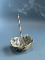 A ceramic incense holder shaped like a leaf with a single incense stick standing inside, set against a soft blue-gray background with wisps of smoke rising.