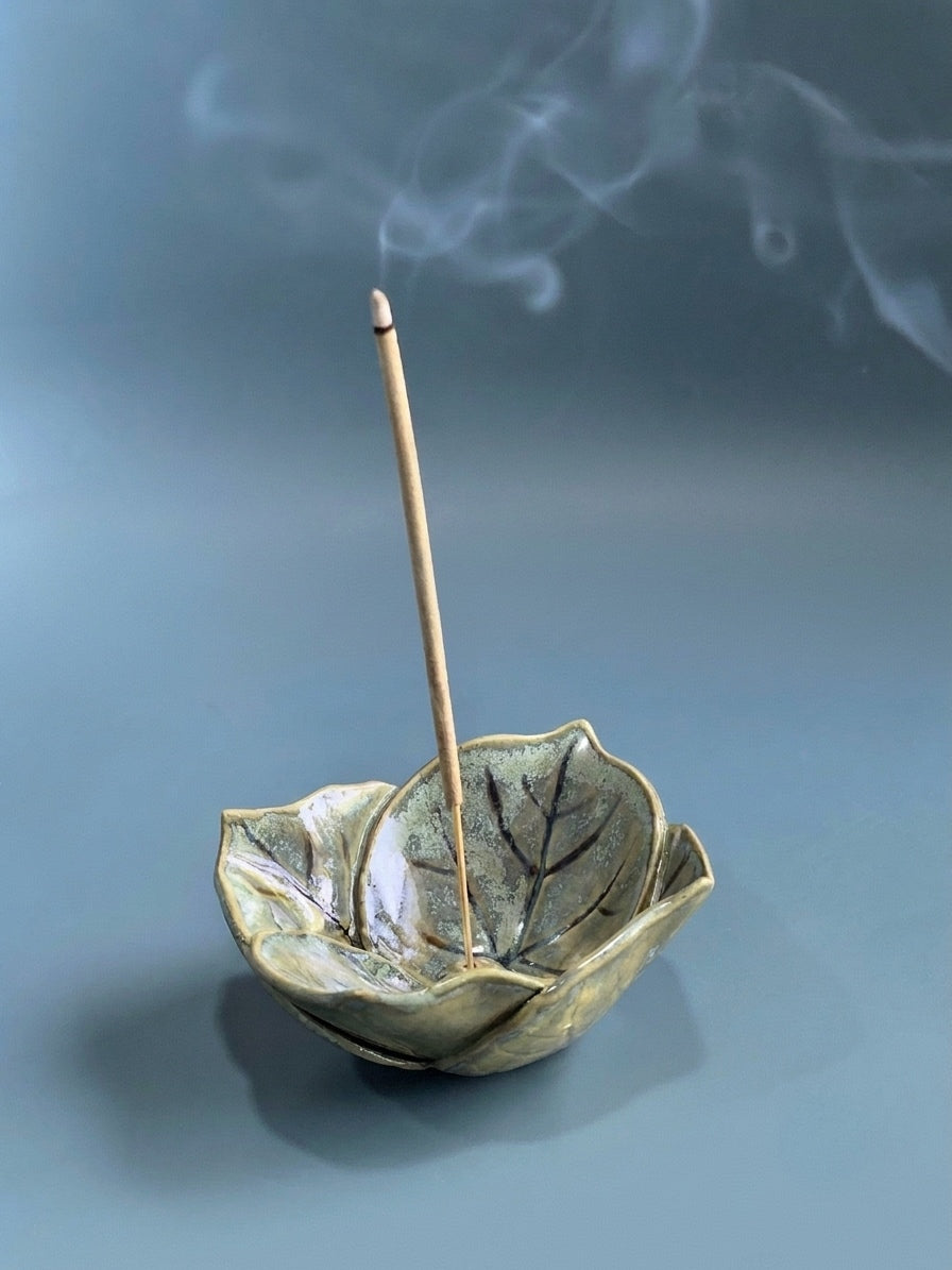 A ceramic incense holder shaped like a leaf with a single incense stick standing inside, set against a soft blue-gray background with wisps of smoke rising.